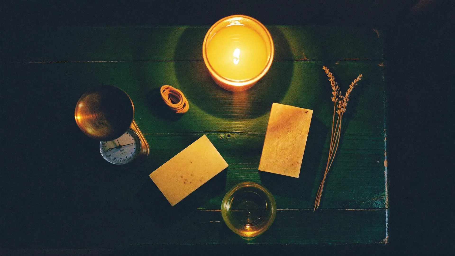 A maker's bench by candlelight: two curing soap bars, a brass scale, lavender and a burning candle jar